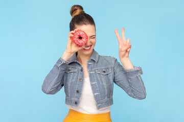 Portrait of funny playful woman with bun hairstyle wearing denim jacket covering mouth with donut showing victory sign. Indoor studio shot isolated on light blue background.