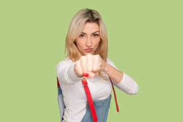 Portrait of serious strict strong adult blond woman clenched fist, boxing, being ready to attack, fighting, wearing denim overalls. Indoor studio shot isolated on light green background