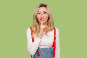 Portrait of cute charming adult blond woman keeps finger near mouth, keeping secret, looking at camera, wearing denim overalls. Indoor studio shot isolated on light green background