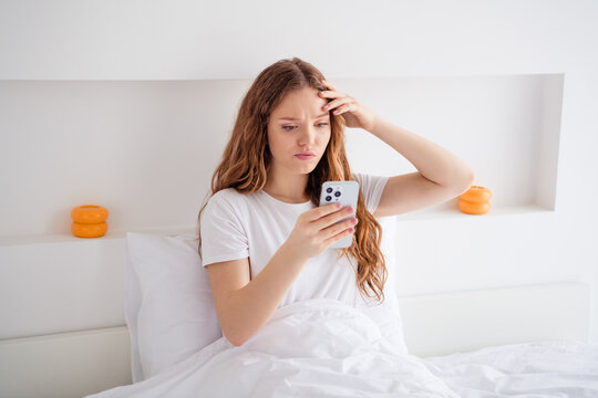 Portrait of female person sit bed under blanket hand touch head use smart phone read news bright bedroom inside