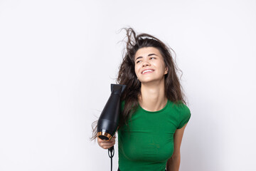 A cute young girl dressed in a green top dries her beautiful long silky hair with a hair dryer against a white background.