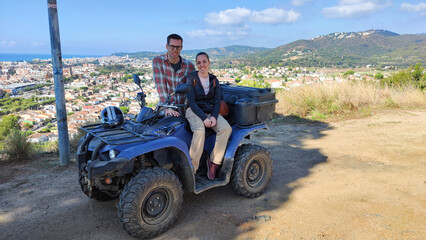 Fototapeta premium Young couple on the top of the mountain with the quad, off-road route through the mountain to the viewpoint of the coastal town. They are posing for the photo with the off-road vehicle.