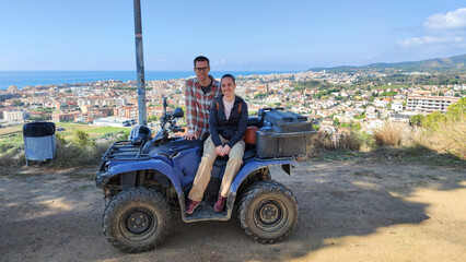 Fototapeta premium Young couple on the top of the mountain with the quad, off-road route through the mountain to the viewpoint of the coastal town. They are posing for the photo with the off-road vehicle. 