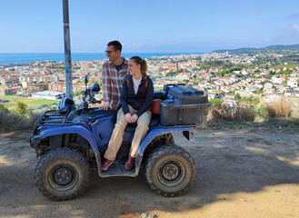 Fototapeta premium Young couple on the top of the mountain with the quad, off-road route through the mountain to the viewpoint of the coastal town. They are posing for the photo with the off-road vehicle.