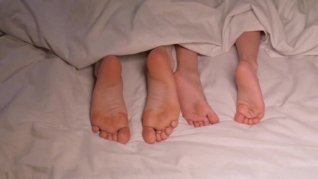 restless legs of the boy son child and the mother female foot lie under the blanket on the sheet in the evening. Baby kid girl daughter and woman mom wiggling bare feet in bed in the bedroom