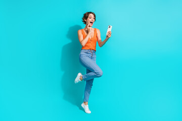 Fototapeta na wymiar Full length photo of overjoyed funky woman wear orange singlet jeans look at eshop in smartphone isolated on turquoise color background
