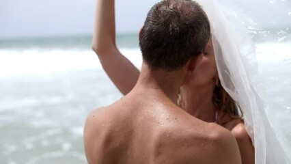 Cheerful newlyweds share tender kiss hiding under bridal veil. Married couple enjoys romantic atmosphere standing in warm ocean water
