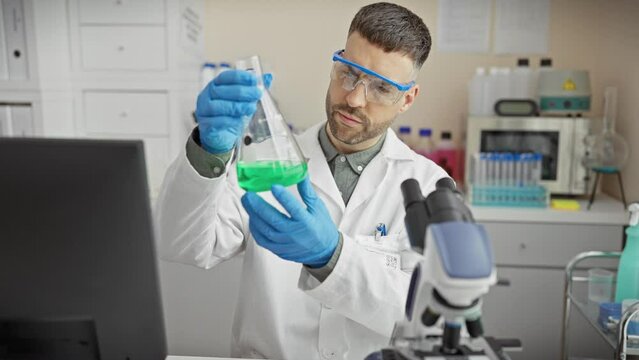 A focused man conducts experiments in a laboratory wearing protective glasses and gloves while examining a green liquid.