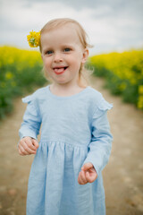 Beautiful girl 3 years old in yellow rubber boots and cute blue dress standing at the yellow flowers field 