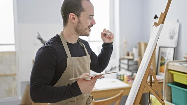 A bearded man paints on a canvas in a bright art studio.