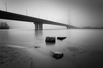Stones in the water stones on the river bank in the foreground and the cable-stayed bridge over the foggy morning river Dnipro. Kyiv. Ukraine. Black and white artistic image on a long exposure. - 769944543
