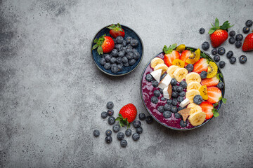 Healthy summer acai smoothie bowl with chia seeds, fresh banana, strawberry, blueberry, cocos, kiwi top view on rustic concrete background with spoon. Space for text