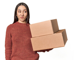 Caucasian young woman with moving boxes on studio background confused, feels doubtful and unsure.