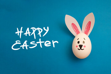 Food photo for Easter holiday. Chicken egg with cute rabbit face and bunny ears on blue background. Greeting card. Happy Easter handwritten white text. Preparation for celebration. Family holiday