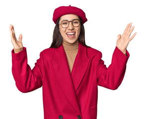 Stylish young Caucasian woman in red trench coat and beret on studio background receiving a pleasant surprise, excited and raising hands.
