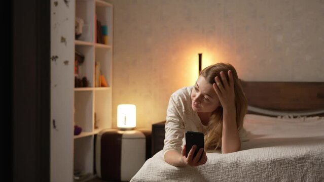 Young blonde woman in pajamas lying on bed speaking over smartphone mobile phone perplexed by the stupid silly interlocutor 