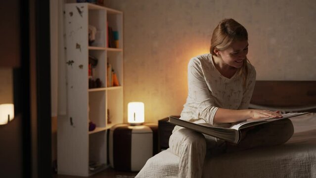 Happy smiling blonde woman in pajamas sitting on bed in cozy comfortable bedroom watching a photo album with photographs