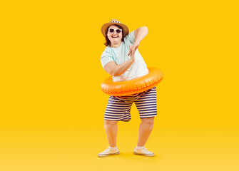 Funny overweight woman dancing with inflatable rubber ring. Happy plus size, fat woman in summer clothes, hat and sunglasses going on summer vacation and having fun over isolated yellow background