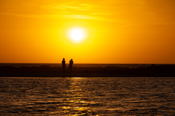 Man and woman staying and looking at sunset on ocean beach, orange sky, silhouettes of people on vacation