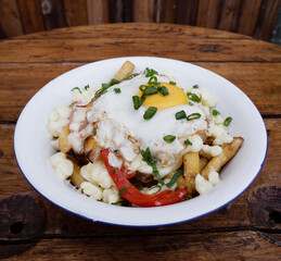 Closeup view of fried potatoes with melted cheese, chorizo, bell pepper, onion and a grilled egg in a white bowl on the wooden table.	