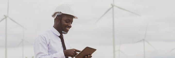 African engineer smile and using tablet with the wind turbine for inspect the operation of large wind turbines that converts wind energy into electrical energy