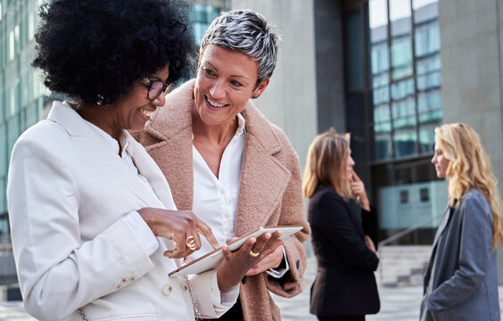 Multiracial mature businesswomen discussing cheerfully the new project ideas together, using the work digital tablet, standing outside financial district. Copy space.