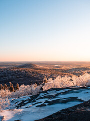 Golden hour above the summit of Mount Watatic in Ashburnham Massachusetts. The mountain and forest is covered in ice and snow, which is reflecting the golden sun. A small mountain and lake are visible