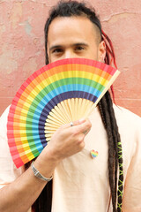 A man with rainbow hair is holding a fan and wearing a heart necklace. Concept of pride and...