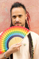 A man with rainbow hair holding a fan. The fan is colorful and has a rainbow pattern. The man is...