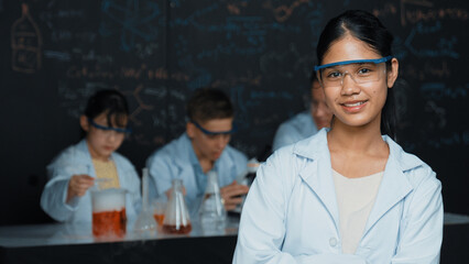 Girl looking at camera with arm folded while people doing experiment at laboratory. Cute student...