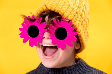 A jubilant young girl sporting playful flower-shaped sunglasses and a yellow beanie laughs heartily against a sunny yellow background
