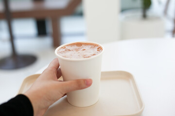 hot cocoa chocolate in paper cup holding by human hand with white warm tone café background 