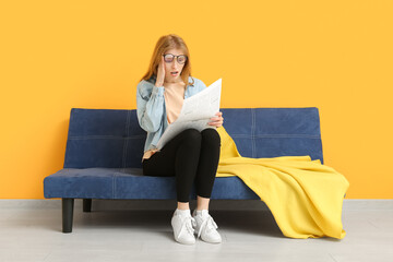 Emotional young woman reading newspaper on sofa near color wall