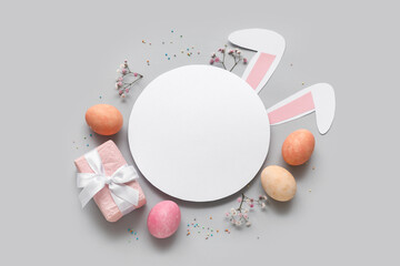 Painted Easter eggs with blank card, gift box and paper bunny ears on grey background