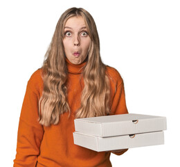 Blonde woman holding pizza boxes in studio shrugs shoulders and open eyes confused.