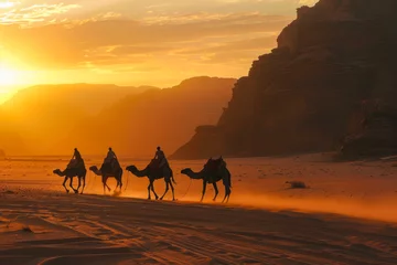 Fototapeten Caravan of camels trekking through a desert at sunset, with the silhouette of rocky mountains in the background © Ilia