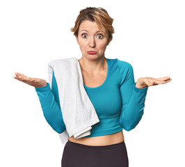 Caucasian woman in sportswear with towel doubting and shrugging shoulders in questioning gesture.