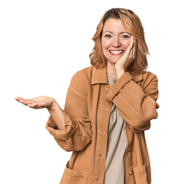 Blonde middle-aged Caucasian woman in studio holds copy space on a palm, keep hand over cheek. Amazed and delighted.