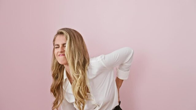 Attractive young blonde woman standing in pain, wearing a shirt over pink background. she's suffering from severe backache, touching aching muscle with hand