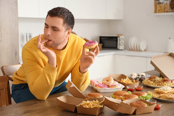 Young man with doughnut licking fingers at table full of unhealthy food in kitchen. Overeating concept