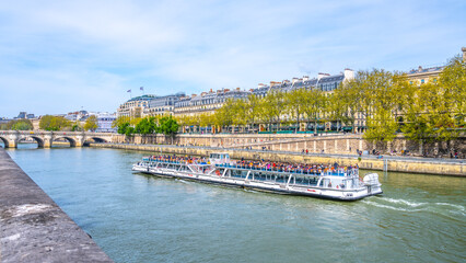 Naklejka premium A riverboat filled with tourists floats along the Seine River in Paris, with city buildings and a bridge in the background. France