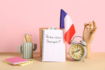 Notebook with French phrase, flag, stationery and alarm clock on table against color background