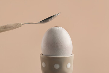 Teaspoon breaking egg in polka dot holder on beige background, closeup