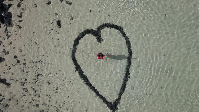 Aerial view of a woman standing inside a heart shape drawn in the sandy bottom of a clear water beach, sunset in Fuerteventura