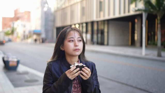 A young Japanese woman from Okinawa Prefecture in her 20s eating Okinawa street food pork egg rice balls on Kokusai Street in Naha City, Okinawa Prefecture 沖縄県那覇市の国際通りで沖縄のストリートフードのポークたまごおにぎりを食べる20代の沖縄
