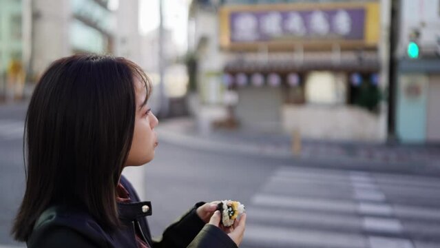 A young Japanese woman from Okinawa Prefecture in her 20s eating Okinawa street food pork egg rice balls on Kokusai Street in Naha City, Okinawa Prefecture 沖縄県那覇市の国際通りで沖縄のストリートフードのポークたまごおにぎりを食べる20代の沖縄