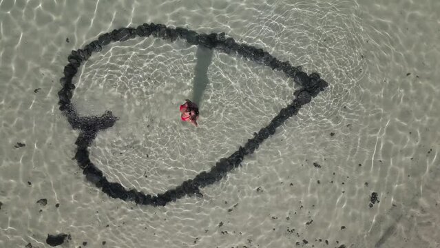 Aerial view of woman standing inside a heart shape drawn in the sandy bottom of a clear water beach, sunset in Fuerteventura
