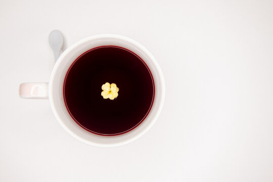 Xícara de chá de Hibisco, vermelho com flor amarela no chá. Tons de vermelho, branco e amarelo, fundo branco. Acidez. Pequena colher branca na alça da xícara. Fundo branco