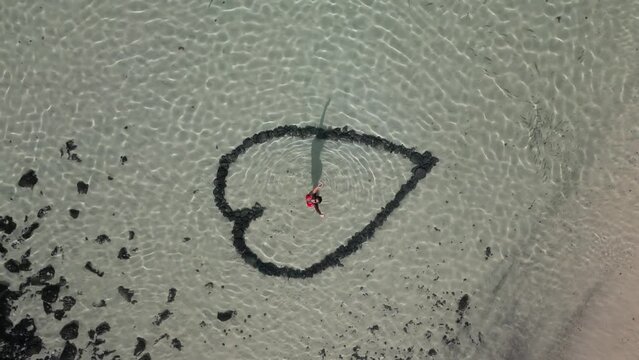 Aerial view of woman standing inside a heart shape drawn in the sandy bottom of a clear water beach, sunset in Fuerteventura