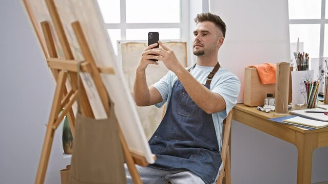 Bearded man in apron using smartphone to take photo of his painting in bright art studio.